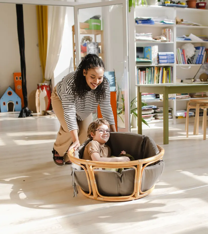 Teacher smiling while pushing a child in a basket seat in a bright playroom